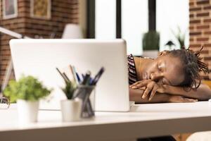 Overworked black businesswoman falls asleep at desk in home office after completing numerous tasks on laptop. Exhausted remote employee naps while seated, recovering from long hours of intense work. photo
