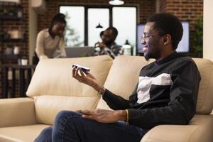 Young businessman seated on couch projects digital content in modern workspace. Behind him, black female manager and team collaborate, reflecting innovation and corporate teamwork. photo