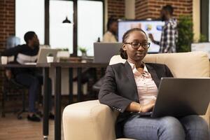 Black female company advisor using laptop, seated on couch, analyzing marketing strategy and compiling reports. African american woman manager comfortably working on business project in startup office photo