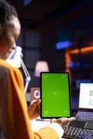 Woman sitting on chair at home office desk in neon illuminated apartment holding green screen tablet while working. Person looking at business graphs on laptop and at mockup device photo