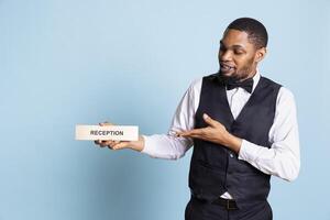 African american hotel porter presenting a wall sign for the reception area, showing an indicator to help guests in finding the front desk upon their arrival at holiday resort. photo