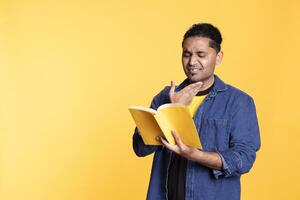 Joyous indian nerd enthusiast reading book for pupils during class, standing against orange background. Happy pleased man holding novel, telling story and relaxing with the lecture in studio. photo