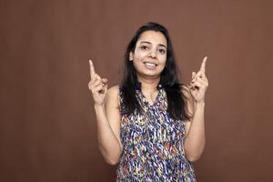 Joyful female model stands against brown background, smiling and pointing upward with both index fingers. Pretty indian woman drawing attention to space above her head for product or ad placement. photo