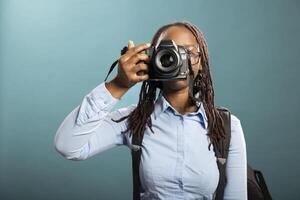 Portrait of black female photographer holding her DSLR, ready for client shoot, showcasing her professionalism. African american woman standing with camera, taking a shot in modern studio. photo