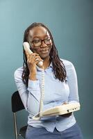 Portrait of smiling black woman using telephone, talking with family and friends in modern studio. African american female model holding landline receiver to her ear, having a phone call. photo