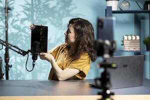Tech specialist presenting tablet in entry level low budget price range to interested viewers, filming with smartphone on tripod. Woman reviewing digital device, recording with mobile phone photo