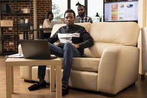 Portrait of african american businessman with glasses, sitting on cozy sofa and looking at camera. Black male employee with laptop on table, seated at lounge area in brick wall office, ready for work. photo