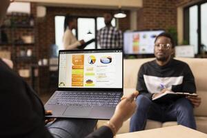 Black manager uses laptop to present business strategies to male employee seated nearby, holding clipboard. African american man on sofa, attentively listens during professional discussion in office. photo