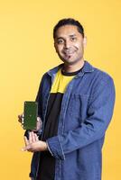 Portrait of indian person doing ad marketing using isolated chroma key on gadget, guy with man bun standing against studio background. Smiling man pointing at empty green screen. photo
