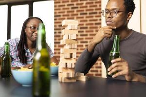 African american man touching his chin, thinking on next move as female friend watches, enjoying jenga game with drinks and snacks. Black guy with beer in hand deciding which wooden block to pull. photo