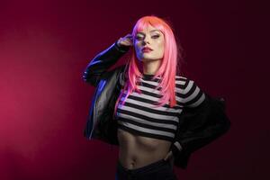 Serious stylish female individual gives striking look while lifting arm in artistic lighting setup in studio. Gorgeous woman with pink hair staring intensely at camera over isolated background. photo