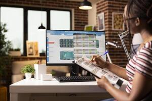 Self employed woman studying financial charts and verifying data using written notes on clipboard. Focused black manager tracks business performance, comparing company data remotely. photo