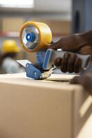 African american man in a logistics center uses duck tape to print invoices, attaching tracking numbers and preparing boxes with AWBs. Operational warehouse supports express delivery. photo