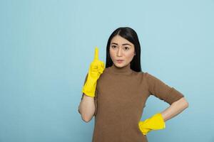 Asian female cleaner in yellow rubber gloves pointing upward, presenting isolated blue background for ads or text. Ideal for cleaning service promotions, banners or marketing visuals. photo