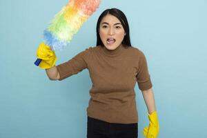 Frustrated asian housekeeper shouting while holding feather duster, overwhelmed by household mess. Angry female janitor screaming toward camera, standing against isolated background. photo