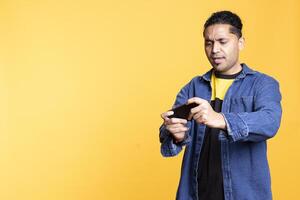 Relaxed Indian guy being entertained by games on mobile phone, enjoying leisure time in studio. Young player having fun playing games and defeating enemies, man bun expression. photo