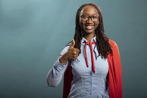 Cheerful black heroine giving a thumbs up, portraying positivity and agreement. African american woman in red cape showing approval gesture and looking at camera with bright smile. photo