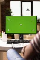 Remote female employee using desktop monitor with green screen mockup. Freelancer seated at desk, analyzing isolated chromakey template for design, editing, or presentation tasks at home. photo