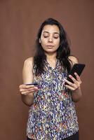 Indian woman using her smartphone to process online payment while holding credit card, standing confidently in studio. Focused female finalizing a transaction on mobile phone, verifying card details. photo