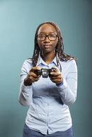 Black woman with glasses holding gaming controller and having fun playing games in modern studio. African american female model in blue shirt fully engaged in a serious gaming session. photo