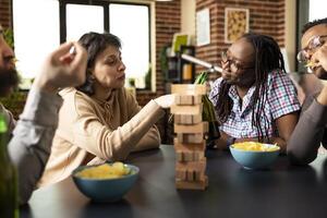 Young women cheer with drinks and guys snack during fun hangout session. Multiethnic group sharing smiles during game break, clinking beer bottles and enjoying chips around table. photo