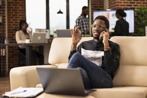 Black male professional in smart casual clothing, sitting on couch and having an important phone conversation with potential client. African american businessman using smartphone in modern workspace. photo