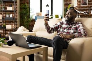 Confident financial advisor seated on cozy sofa, holding cellphone during remote call with client. Male entrepreneur manages virtual meeting with employee about startup goals from living room. photo