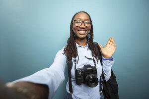 Black female vlogger with dslr around neck waves at camera, recording content in blue studio setting for her channel. African American photographer greets viewers with smile and hello hand gesture. photo