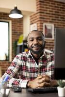 Smiling black remote worker sitting at desk with desktop pc in cozy brick wall living space. Self employed male professional prepared for virtual meetings and business tasks from home. photo