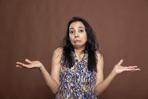 Portrait of indian woman shrugging shoulders with puzzled expression, standing against brown background. Beautiful female model holding hands out, clearly unsure and making I dont know gesture. photo