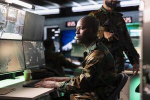 Army unit working in governmental facility operating as surveillance monitoring center filled with digital equipment. African american man doing job in base of operations photo