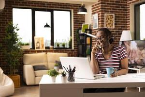 Cheerful black woman reads email on laptop and shares exciting news with colleague during phone call. Remote female manager sits at desk with coffee cup in hand, talking on cellphone while working. photo
