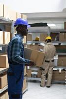 African american worker in protective gear checks merchandise for dispatch in a busy warehouse. Crates and boxes arranged for logistics and transportation in operational supply chain hub. photo