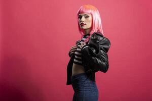 Stylish fashion model wearing striped crop top and leather jacket, glowing with beauty while staring directly at camera with focused gaze. Beautiful carefree woman stands isolated over pink background photo