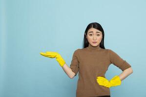 Asian housemaid wearing yellow rubber gloves, pointing to left with open hand gesture. Female cleaner standing against blue background, ideal for housekeeping or cleaning service ads. photo