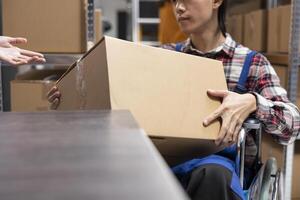 Asian depot employee in wheelchair works at the packaging station with multiple boxes, preparing the orders to be shipped for local clients. Disability friendly startup space. Close up. photo