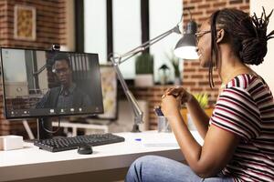 Smiling black woman seated at desk, participates in virtual meeting with potential client. African american female employee working from home, having online discussion with male manager on call. photo