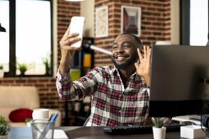 Black male entrepreneur seated at desk with cellphone, happily greeting business partner with wave on call. African american freelancer recording vlog, making hello gesture for online viewers. photo