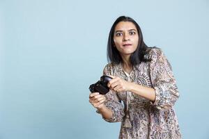 Happy indian woman entertained by games on her console, enjoying leisure time. Player enjoying game on controller, having fun defeating enemies, standing against studio background. photo