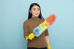 Confident asian female cleaner in yellow rubber gloves holding feather duster, smiling while ready to tackle dust. Focused woman standing against blue backdrop with cleaning tool in hand. photo