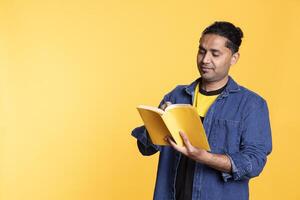 Young man book lover absorbed in a fantasy book against studio background, relaxing with a joyful story. Indian person enjoying lecture in front of the camera, reading enthusiast nerd. photo