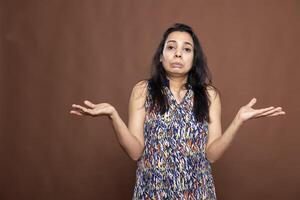 Confused indian lady with pursed lips and shrugged shoulders, expressing uncertainty and ignorance. Pretty female individual standing and looking unsure against isolated background. photo
