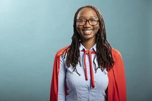 Portrait of black female individual cheerfully standing isolated over blue background. Smiling young lady with glasses and red superhero cape, looking at camera, portraying positivity in modern studio photo