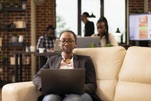 Black female manager sits on cozy couch, using laptop to analyze startup research data and manage company reports. Focused businesswoman with glasses, reviewing emails and preparing business projects. photo