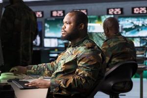 Military early warning systems operator in control room monitoring incoming enemy aerial attacks. African american army agent maintains air space superiority with radar during armed conflict photo