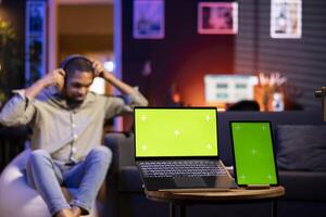 Young black person enjoys music on wireless headset next to isolated display, feeling happy and casual laying on the bean bag in the living room. Relaxed chill man in his trendy loft. photo