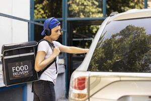Confident woman delivering takeout with the car using a backpack, arriving at the entrance of a modern building. Employee preparing to ring the door bell to bring the food order to client. photo