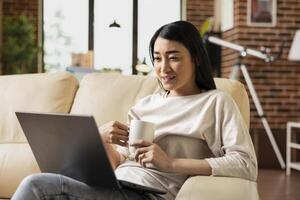 Female freelancer sits with laptop on laps and holds coffee mug, focused on online webinar. Remote asian employee drinking tea while improving business skills in cozy brick wall home. photo