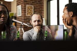 Concerned white man watching emotional content with african american friends in brick wall room. Bearded male individual sitting at table, appears distressed while viewing sad with covered mouth photo