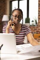 Concerned businesswoman talks on phone and reviews printed financial reports, sitting at messy desk filled with paperwork. Closeup of female freelancer holding document and speaking on mobile device. photo